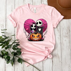 Puede incluir: Camiseta rosa claro con un gr&aacute;fico de una pareja y una calabaza de Halloween dentro de un coraz&oacute;n rosa. La pareja tiene una apariencia esquel&eacute;tica. La calabaza es naranja con una cara tallada. La camiseta es de material suave.