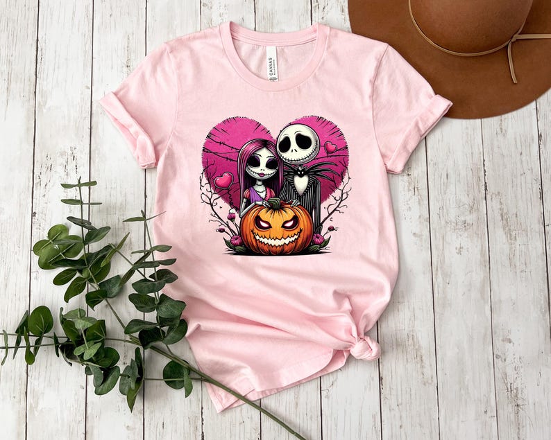 Puede incluir: Camiseta rosa claro con un gr&aacute;fico de una pareja y una calabaza de Halloween dentro de un coraz&oacute;n rosa. La pareja tiene una apariencia esquel&eacute;tica. La calabaza es naranja con una cara tallada. La camiseta es de material suave.