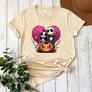 Puede incluir: Camiseta beige con un gr&aacute;fico colorido de Jack Skellington y Sally de The Nightmare Before Christmas, enmarcado por un coraz&oacute;n rosa y una calabaza tallada. El dise&ntilde;o incluye a Jack, Sally y una calabaza naranja sonriente.