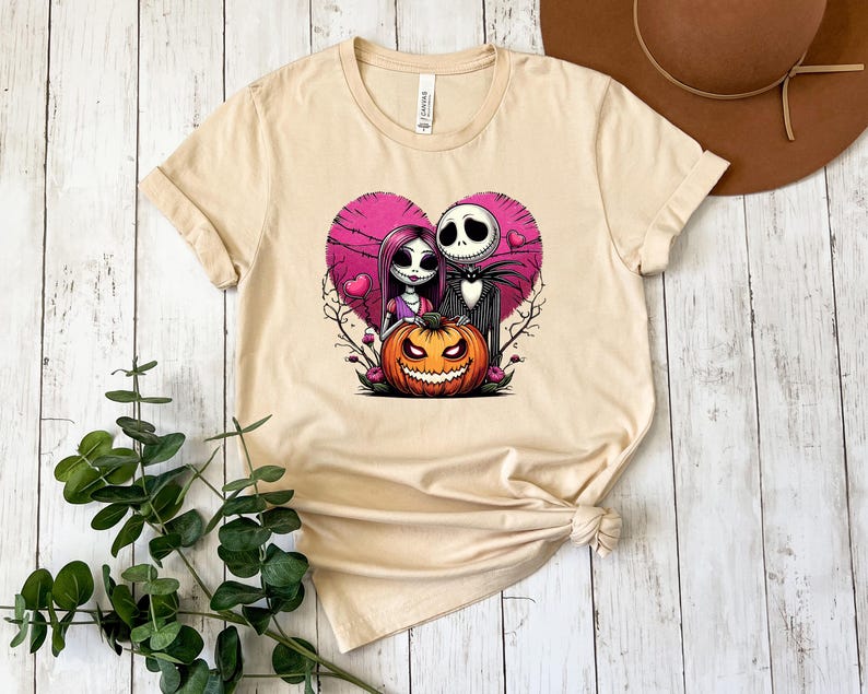 Puede incluir: Camiseta beige con un gr&aacute;fico colorido de Jack Skellington y Sally de The Nightmare Before Christmas, enmarcado por un coraz&oacute;n rosa y una calabaza tallada. El dise&ntilde;o incluye a Jack, Sally y una calabaza naranja sonriente.