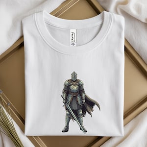 May include: White t-shirt featuring a detailed illustration of a medieval knight in full armor, holding a sword. The knight is depicted in shades of gray, silver, and brown. The shirt is folded neatly on a gold tray.