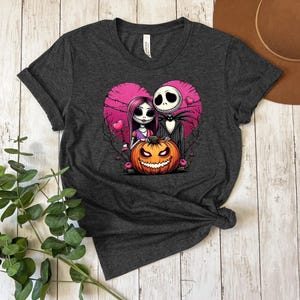Puede incluir: Camiseta gris oscuro con un dise&ntilde;o de Jack y Sally de Pesadilla antes de Navidad dentro de un coraz&oacute;n rosa. El dise&ntilde;o incluye una calabaza tallada. La imagen de estilo caricatura est&aacute; sobre una superficie de madera.