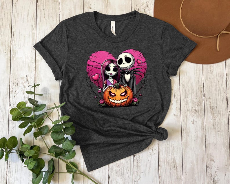 Puede incluir: Camiseta gris oscuro con un dise&ntilde;o de Jack y Sally de Pesadilla antes de Navidad dentro de un coraz&oacute;n rosa. El dise&ntilde;o incluye una calabaza tallada. La imagen de estilo caricatura est&aacute; sobre una superficie de madera.