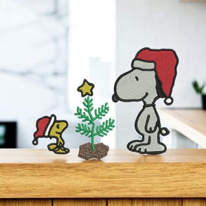 May include: Embroidered Christmas-themed Snoopy and Woodstock characters with a small Christmas tree. Snoopy and Woodstock are wearing red Santa hats. The tree has a gold star on top and is on a wooden surface.