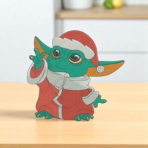Op de afbeelding: Geborduurde figuur van een groen personage met een rode kerstmuts en gewaad met witte rand. Het personage heeft grote ogen en zwaait. De figuur staat op een lichtbruin oppervlak.