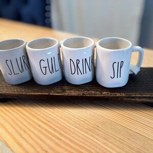 May include: Four white ceramic mugs with black text reading "SLUR", "GUL", "DRINK", and "SIP" on a dark wooden tray. The mugs have small handles and are arranged on a rectangular tray, ideal for serving drinks.