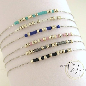 May include: A collection of six delicate silver-tone chain bracelets, each featuring a unique sequence of metallic silver beads interspersed with colored beads in turquoise, white, blue, pink, grey, and black. The logo Devin Michaels is visible.