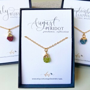 May include: Three small boxes with gold necklaces and gemstone pendants. The boxes are labeled with the months of July, August, and September, and the corresponding birthstones: Ruby, Peridot, and Sapphire. Each box also includes the stone's associated qualities.
