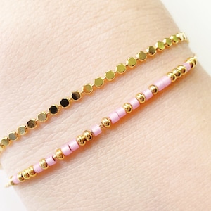 May include: Two gold bracelets on a wrist. One bracelet is made of small gold beads and the other is made of larger gold beads.