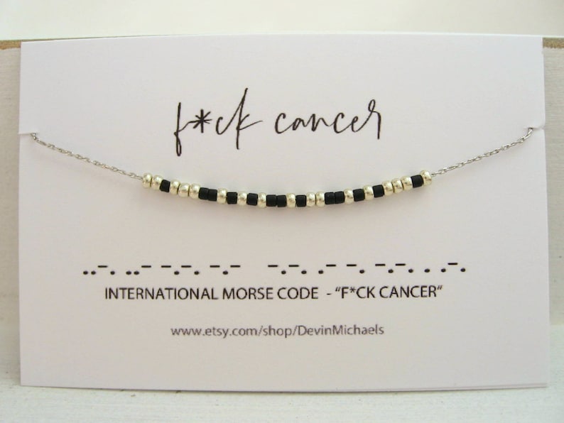 May include: A silver necklace with black and silver beads spelling out "F*ck Cancer" in Morse code. The necklace is displayed on a white card with the phrase "f*ck cancer" written above the necklace.