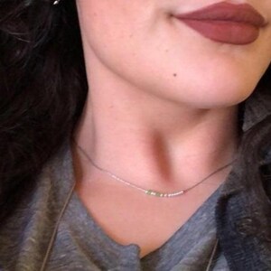 May include: A delicate silver necklace with small beads and a few green stones. The necklace is worn against a gray top, with a person's lips visible in the background, painted with a dark red lipstick.