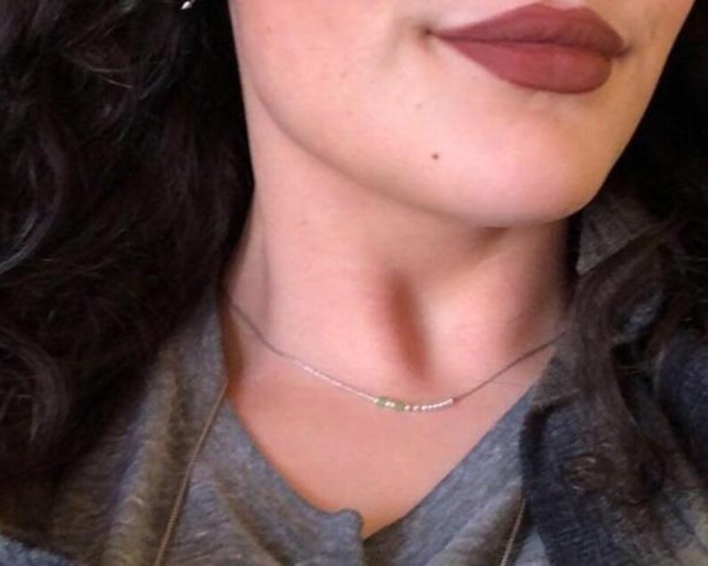 May include: A delicate silver necklace with small beads and a few green stones. The necklace is worn against a gray top, with a person's lips visible in the background, painted with a dark red lipstick.