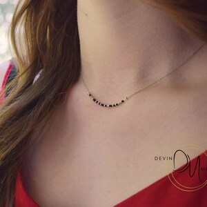 May include: A delicate necklace with a thin silver chain. The necklace features a series of small, black beads interspersed with gold-colored beads, forming a gentle curve across the neckline. The background is blurred, with a red top visible.