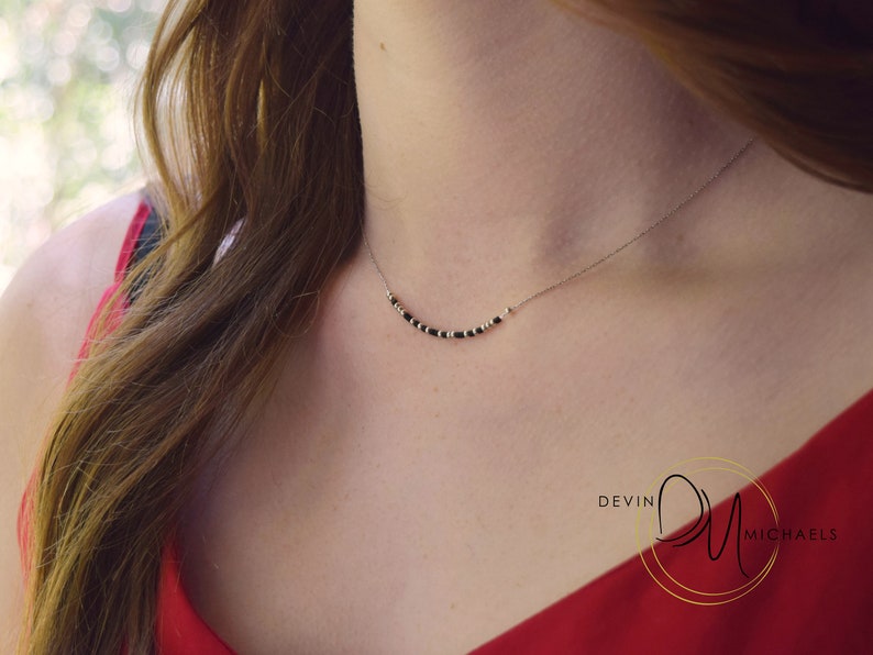 May include: A delicate necklace with a thin silver chain. The necklace features a series of small, black beads interspersed with gold-colored beads, forming a gentle curve across the neckline. The background is blurred, with a red top visible.
