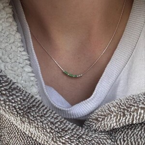 May include: A delicate silver necklace featuring a row of small, light green beads. The beads are strung along a thin silver chain. The necklace is shown against a soft, neutral background, highlighting the jewelry's simple elegance.
