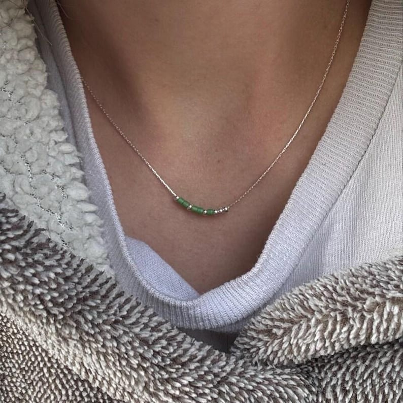 May include: A delicate silver necklace featuring a row of small, light green beads. The beads are strung along a thin silver chain. The necklace is shown against a soft, neutral background, highlighting the jewelry's simple elegance.
