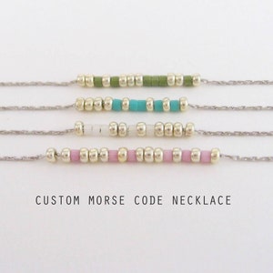 May include: Four silver chains with different colored beads, including pink, white, turquoise, and green. The chains are hanging from a white background. The text "CUSTOM MORSE CODE NECKLACE" is below the chains.