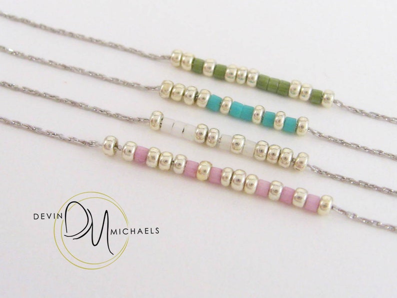 May include: Four delicate necklaces with silver chains and colorful beaded accents. The beads are in shades of green, turquoise, white, and pink. The necklaces are displayed on a white background. The brand logo "Devin Michaels" is in the lower left corner.
