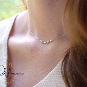 May include: A delicate silver necklace with small, colorful beads. The necklace is worn on a person's neck, with the beads resting near the collarbone. The background is blurred, suggesting an outdoor setting. The necklace is a piece of jewelry.