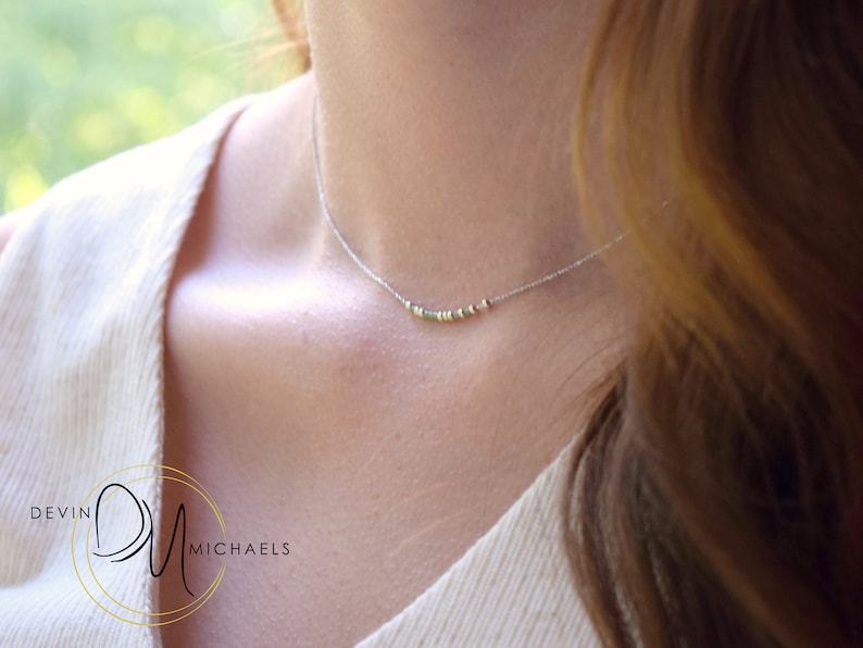 May include: A delicate silver necklace with small, colorful beads. The necklace is worn on a person's neck, with the beads resting near the collarbone. The background is blurred, suggesting an outdoor setting. The necklace is a piece of jewelry.