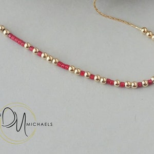 May include: A delicate gold necklace featuring alternating gold beads and small red beads. The necklace is displayed on a light gray surface. The brand name "Devin Michaels" is visible in the lower left corner.
