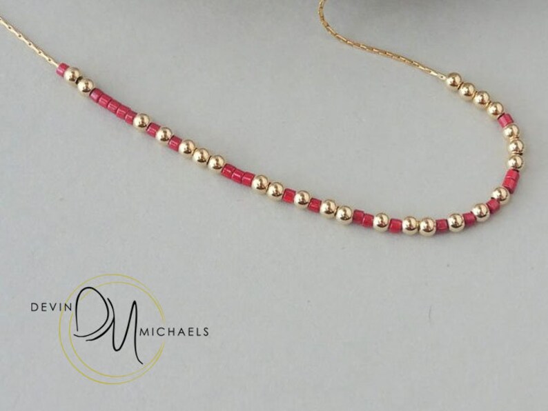 May include: A delicate gold necklace featuring alternating gold beads and small red beads. The necklace is displayed on a light gray surface. The brand name "Devin Michaels" is visible in the lower left corner.