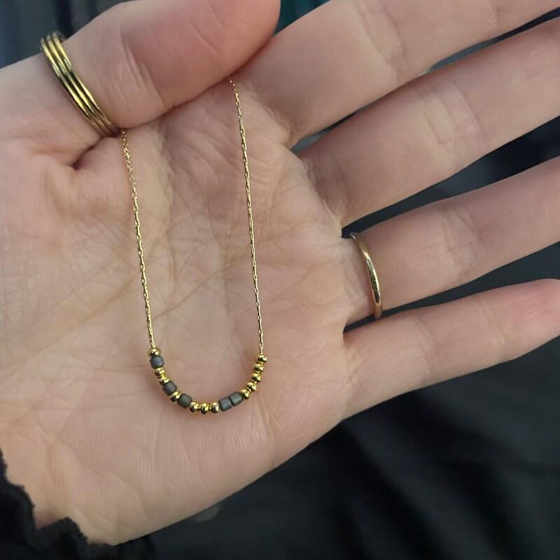 May include: A delicate gold necklace featuring a series of small, dark gray beads interspersed with smaller gold beads. The necklace is displayed against a neutral background, highlighting its simple elegance. The necklace is a fine piece of jewelry.