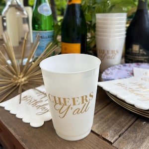 May include: A white plastic cup with the text "Cheers Y'all" in gold lettering. The cup is on a wooden table, with other party supplies like napkins and plates. Bottles of champagne and sparkling water are in the background.
