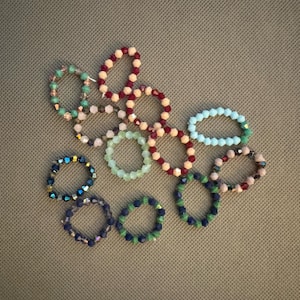 May include: An assortment of beaded rings in various colors, including teal, pink, blue, and green, are arranged on a textured, neutral-toned surface. The rings feature faceted beads, creating a sparkling effect. The collection showcases a variety of styles and color combinations.