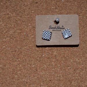 Puede incluir: Pendientes cuadrados con estampado de cuadros blancos y negros. Se exhiben en una pequeña etiqueta de cartón marrón con el texto "Sarah Shinee Creations". La etiqueta está fijada a una superficie de corcho marrón.