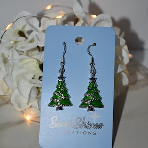 May include: A pair of silver-toned hook earrings featuring green enameled Christmas tree charms. The earrings are attached to a light blue display card with the text Sarah Shines CREATIONS printed in white at the bottom of the card.