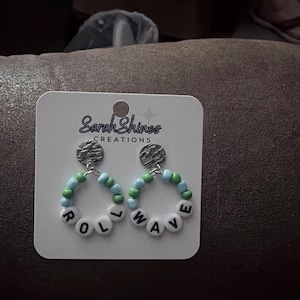 May include: A pair of earrings on a white card with the text "Sarah Shines CREATIONS". The earrings have a silver top with a beaded hoop design. The hoops are blue, green, and white, spelling out the word "ROLL WAVE".