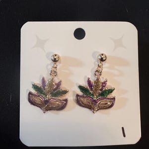 May include: A pair of gold-toned earrings featuring a Mardi Gras mask design. The earrings have a gold ball stud and a dangling mask with purple, green, and gold enamel details. They are displayed on a white card.