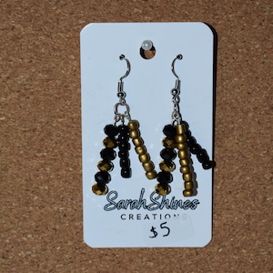 May include: A pair of dangle earrings with black and gold beads arranged in an abstract design. The earrings are displayed on a white card with the text "Sarah Shines Creations". The background is a textured, brown surface.