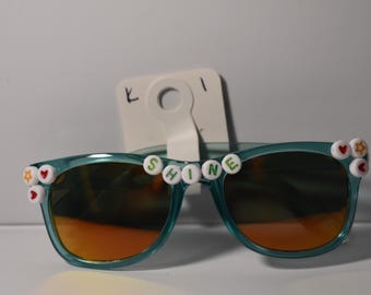 May include: Teal sunglasses with orange lenses. The word "SHINE" is spelled out across the top with white beads. The frames are decorated with heart and star-shaped embellishments. A white tag is attached.