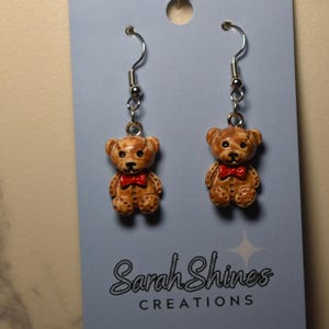 May include: A pair of teddy bear earrings. Each brown bear has a red bow tie and is suspended from a silver-colored hook. The earrings are displayed on a light blue card with the text "Sarah Shines Creations".