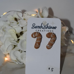 Puede incluir: Un par de pendientes en forma de bastón de caramelo sobre una tarjeta blanca con el texto "Sarah Shines CREATIONS". Los pendientes son marrones con rayas de glaseado blanco.