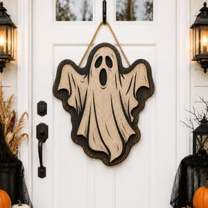 Puede incluir: Un adorno de pared de madera con forma de fantasma, compuesto por una silueta de color beige claro con acabado desgastado y recortes negros para los ojos y la boca, montado sobre un fondo marrón oscuro y colgado con una cuerda de yute gruesa.