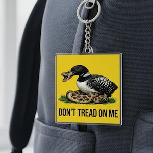 Può includere: Portachiavi quadrato con sfondo giallo. Presenta un uccello colimbo che morde un serpente, con la scritta "DON'T TREAD ON ME". L'uccello è nero e bianco, e il serpente è marrone e beige. Il portachiavi è attaccato a una catena d'argento.