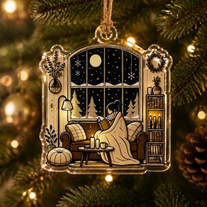 May include: A clear acrylic Christmas ornament featuring a printed illustration of a cozy indoor scene with a person reading on a sofa by a window, surrounded by a bookshelf, a floor lamp, and candles, hanging from a rustic twine loop.
