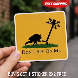 May include: A square sticker with a yellow background featuring a black silhouette of a raccoon sawing down a utility pole topped with security cameras. Below the raccoon, the text reads DON'T SPY ON ME in black, bold, serif capital letters.