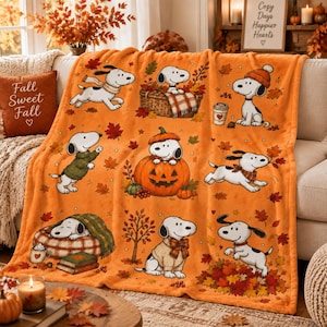 May include: An orange fleece throw blanket featuring a repeating pattern of Snoopy in various autumn-themed scenes, including sitting in a pumpkin, wearing a sweater, and playing in fallen leaves, accented with scattered red and yellow maple leaves.