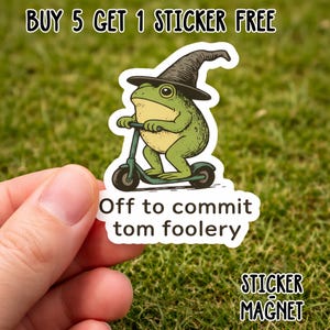May include: A sticker with a cartoon frog in a witch's hat on a scooter. The frog is green and tan, with a black hat. Text on the sticker says "Off to commit tom foolery" and "STICKER MAGNET".