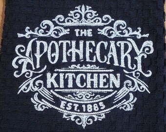 May include: Black kitchen towel with white embroidery that reads "The Apothecary Kitchen Est. 1885".