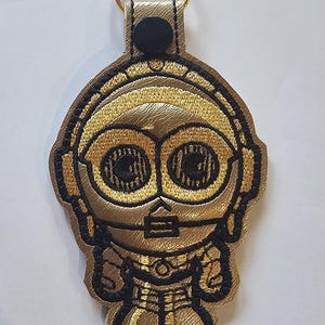 May include: Gold and black embroidered keychain featuring a stylized C-3PO design. The keychain has a gold-colored metal ring and a black snap closure. The design is detailed with black stitching on a gold background.