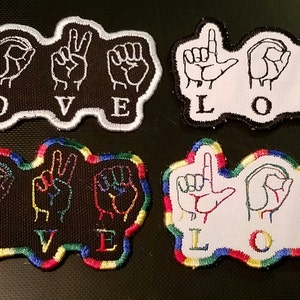 May include: Four embroidered patches with the word "LOVE" spelled out in American Sign Language. The patches are in two different styles: one with a white outline and the other with a rainbow outline. The patches are on a black background.