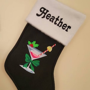 May include: Black Christmas stocking with a white cuff that says "Heather". The stocking features an embroidered martini glass with a green bow, a red and white straw, and a lemon wedge.