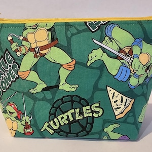 May include: Green fabric cosmetic bag featuring Teenage Mutant Ninja Turtles illustrations. The bag has a yellow zipper and is patterned with the turtles, pizza slices, and the words "Turtle Power" and "Turtles".