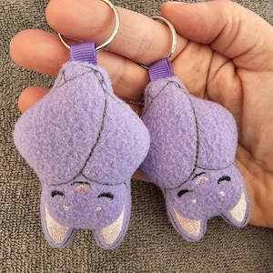 Op de afbeelding: Twee paarse pluche vleermuis sleutelhangers met geborduurde ogen en oren. De sleutelhangers hangen aan zilveren sleutelringen.