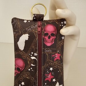 May include: A dark brown tarot card-themed keychain wallet with a gold ring. The wallet features a zipper and illustrations of skulls, stars, and tarot card imagery. The text "The Moon", "Death", and "The Cat" are visible.